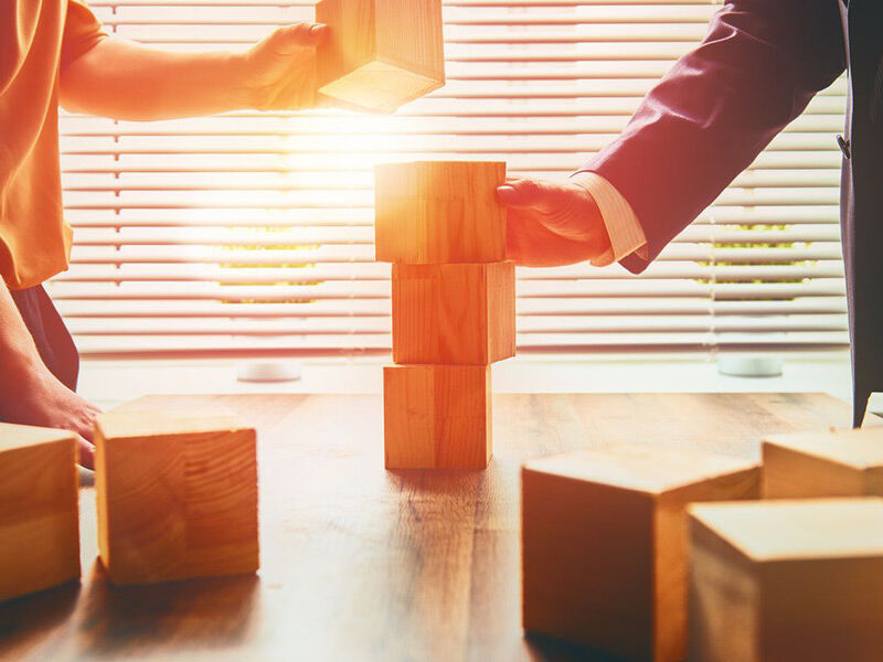 Hand stacking wooden blocks in sunlight, symbolizing teamwork and strategy.