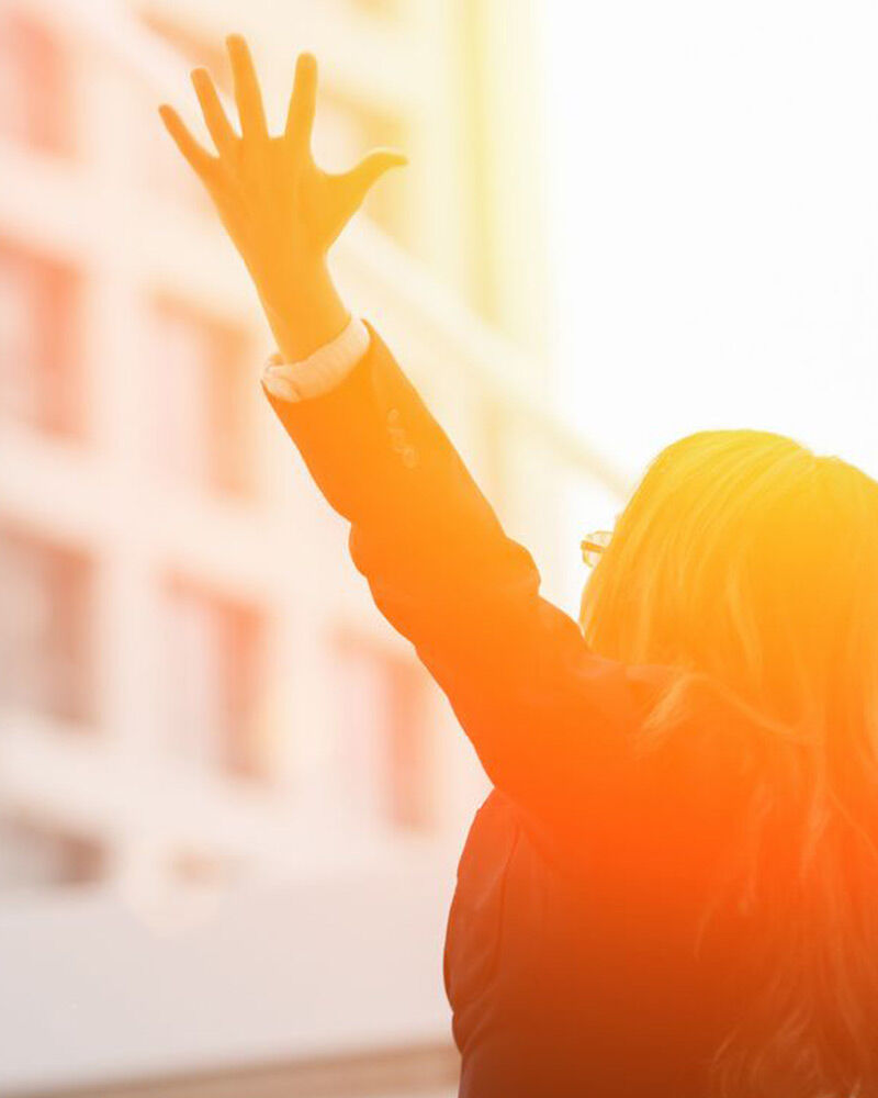 Person raising hand toward bright sunlight in an urban setting.
